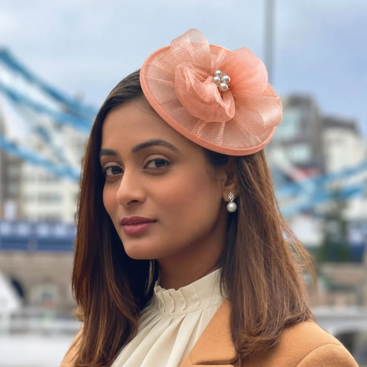 Woman wearing the Elaria Vintage Peach Fascinator with sculpted mesh loops and pearl centre near Tower Bridge.